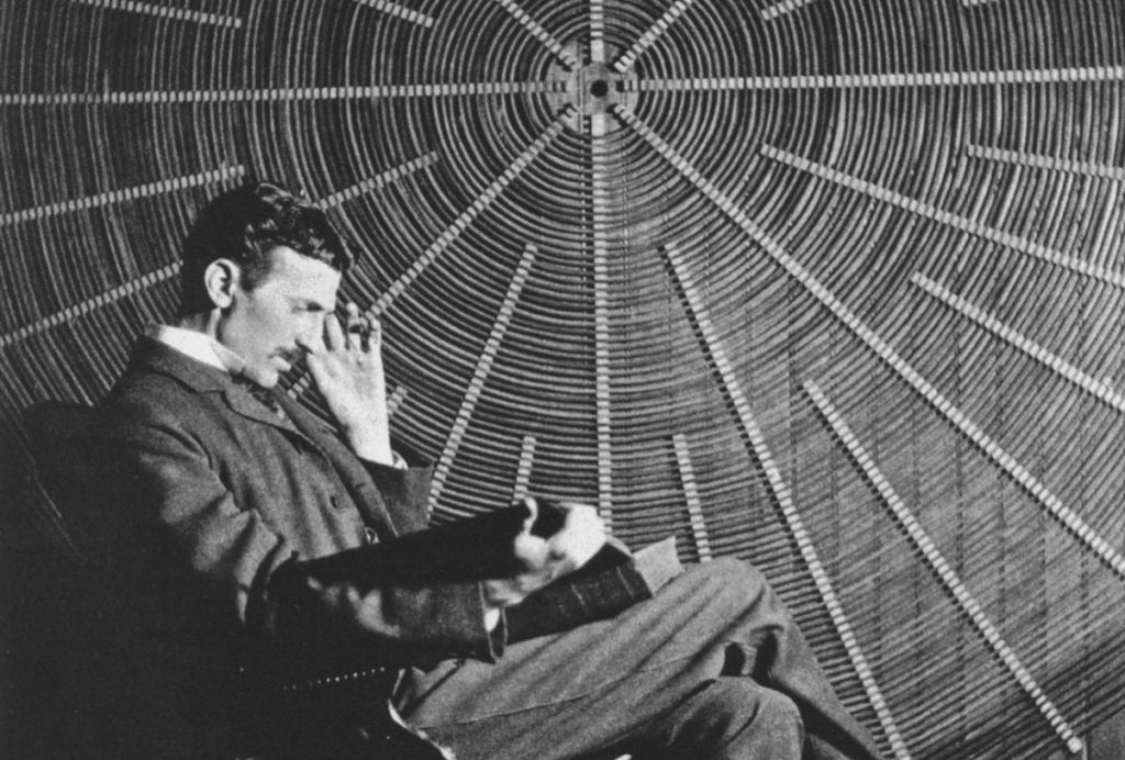 Tesla sitting in front of a spiral coil used in his wireless power experiments at his East Houston St. laboratory.